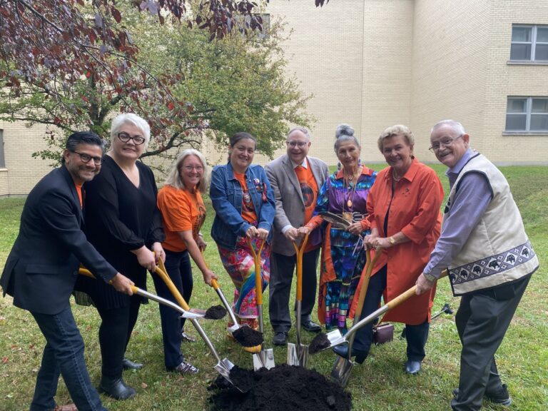 Guests in orange and dark clothing pose with golden ceremonial shovels around a mound of earth at the Chantal Beauvais Courtyard groundbreaking at Saint Paul University.