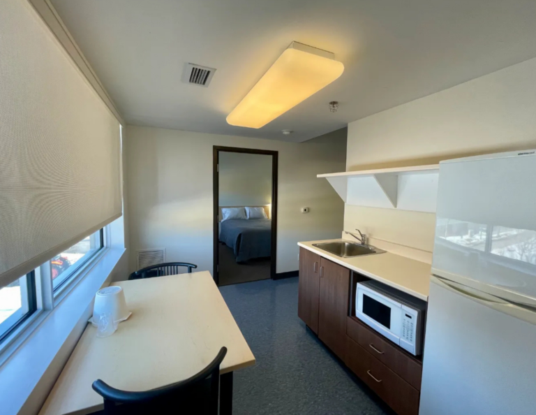 Suite kitchenette with a sink, microwave and fridge, a small table by the window and the bedroom visible through the doorway